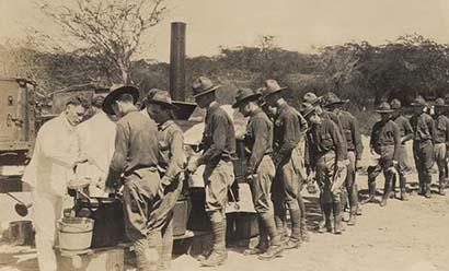 Sepia photograph of several soldiers serving food to a long line of soldiers in an open field.
