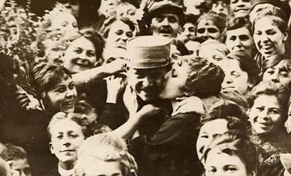 Sepia photograph of a crowd of smiling faces. In the center a woman kisses the cheek of a man in military uniform.