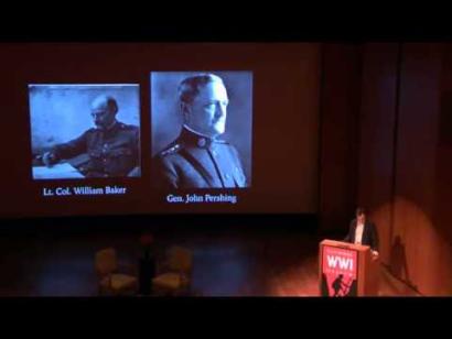Video still of a man standing at a podium on the Museum auditorium stage. Two photographs of WWI generals are on the screen behind the speaker.