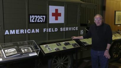 Video still of an older white man, facing the viewer, standing in front of a museum display of a WWI-era ambulance.