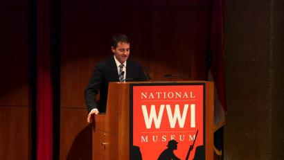 Video still of a white man in a black suit speaking at a podium on the Museum auditorium stage.