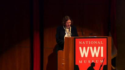 Video still of a white woman speaking at the podium on the Museum auditorium stage.