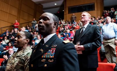 Modern photograph of a filled auditorium. In the foreground, a Black man and a Black woman in military uniform stand and sing.