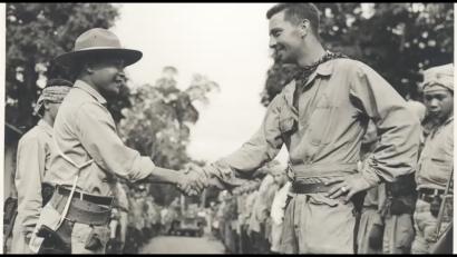 Black and white photograph of two men shaking hands amidst a group of soldiers, one darker-skinned and one lighter-skinned.