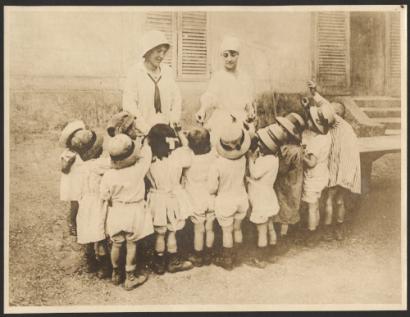 Sepia photograph of a group of children gathered around two women dressed in white.