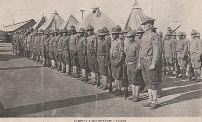 Black and white photograph of two rows of Native American soldiers standing at attention in front of tents and barracks.