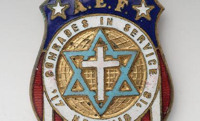 Modern photograph of a small charm shaped like a shield in the colors of the U.S. flag. In the center a cross is inside a star of David. Text engraved around the cross and star: "Comrades in Service / '17 '18 /  Neh. 4:18"