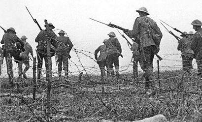 Black and white grainy photograph of WWI soldiers stepping over barbed wire in an open field, all carrying rifles.