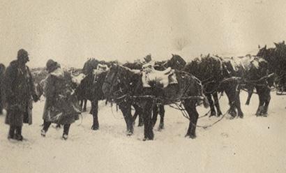Black and white photograph of two soldiers in thick winter coats and a team of horses harnessed to artillery, all covered in snow