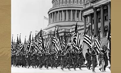 Black and white photograph of men in uniforms and carrying U.S. flags marching in front of the U.S. Capitol building.