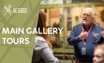 An older white man with a white beard in a blue shirt speaks and gestures while several museum guests listen