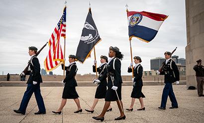 Modern photo of six young people in military uniform marching in a line carrying flags, rifles or swords