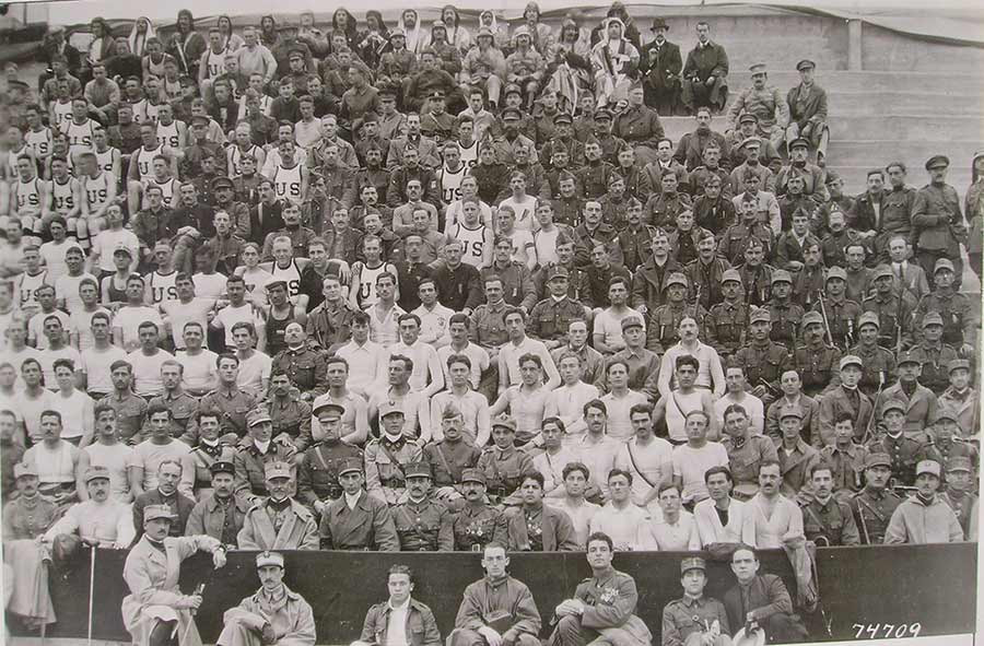 Black and white photograph of a section of the stands in a stadium. The people in the seats are dressed in the sports uniforms for various countries, grouped together by uniform.
