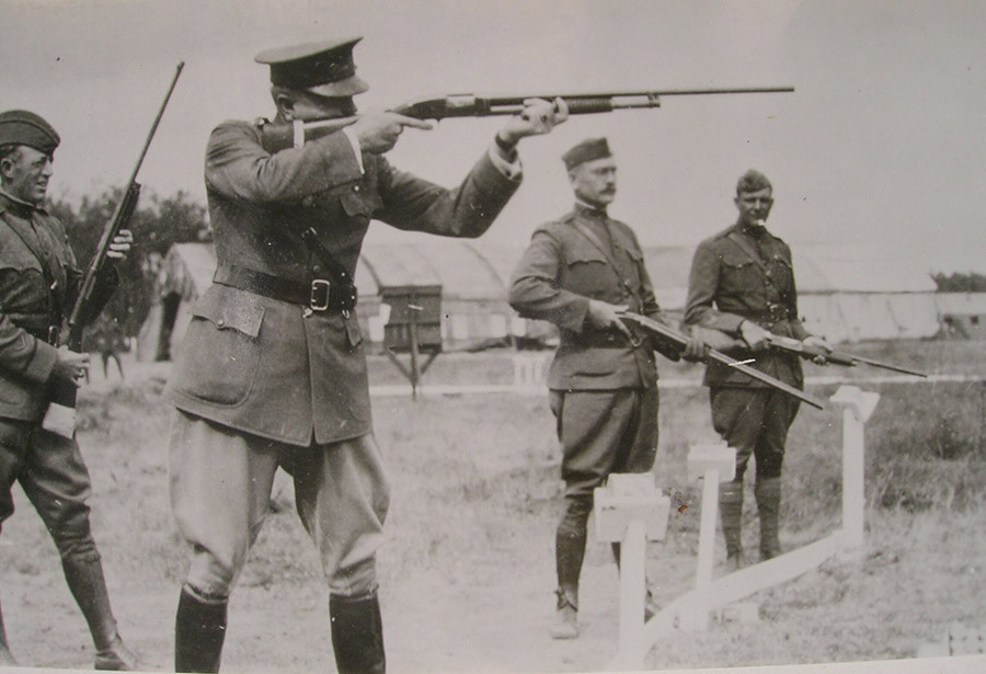 Black and white photograph of four men in military uniform holding rifles. The one in the foreground has his rifle up shooting at an unseen target. The other three are observing.