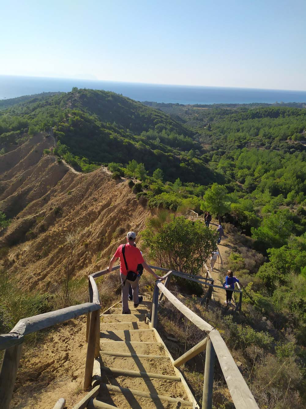 View down a tall ridge that snakes into the distance. Several people walk along a trail built on the top of the ridge. Greenery on both sides.