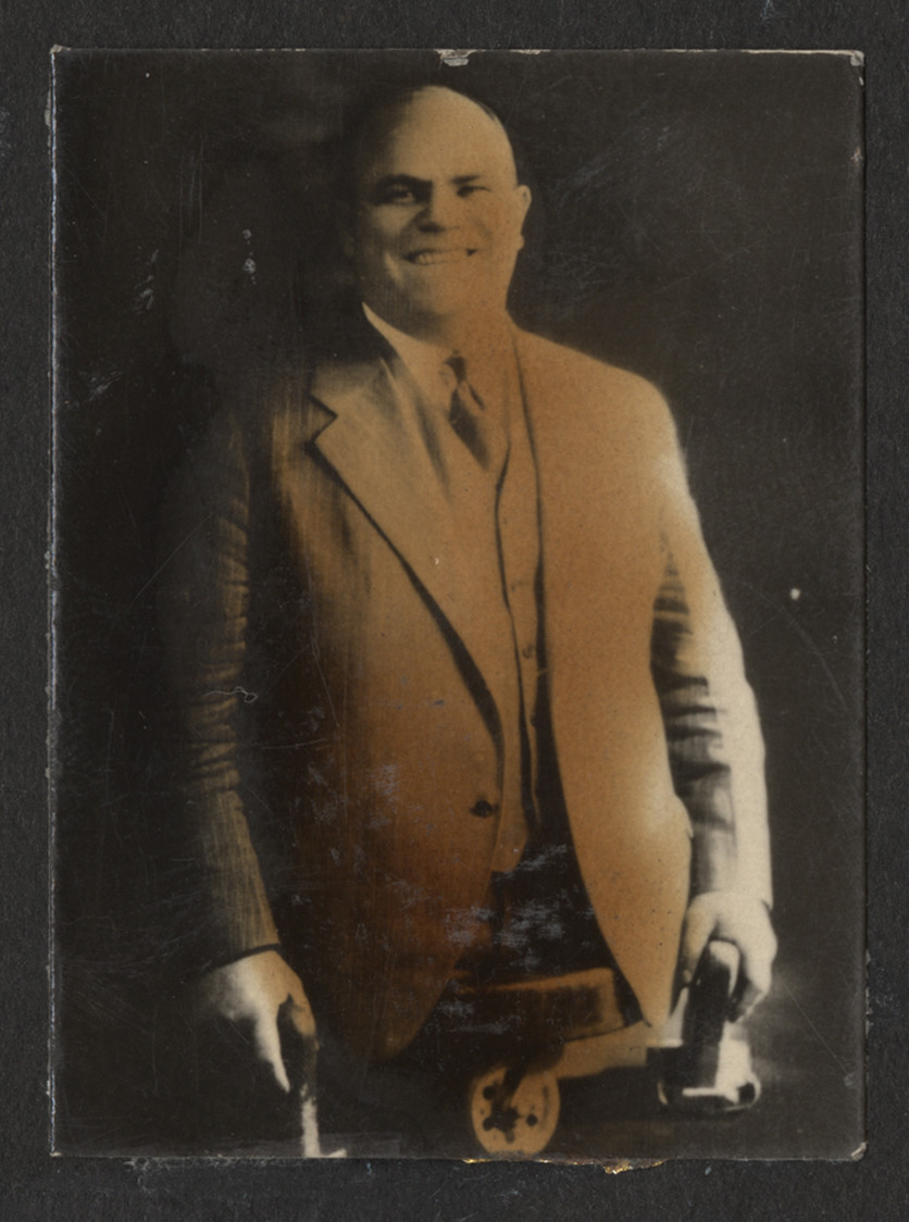 Black and white portrait photograph of a veteran wearing civilian clothing. Man is missing both legs from the hips down. He sits on a flat, three-wheeled cart and holds blocks with handles to help him move. 