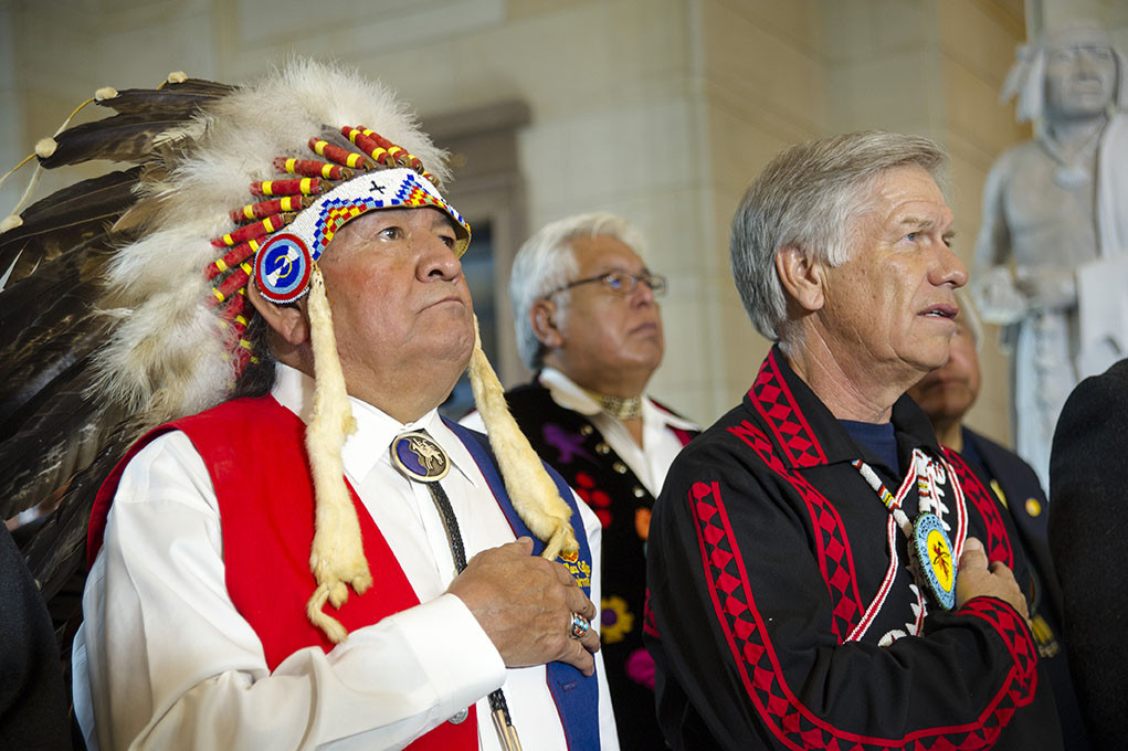 Modern photograph of several older Native American men in formal clothes and regalia, looking forward with their hands on their hearts.