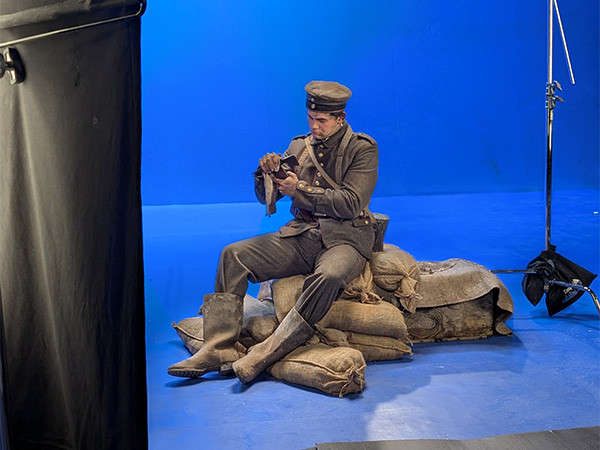 Modern photo of a white male actor in WWI military uniform sitting on a pile of sandbags in a bluescreened studio, surrounded by lights and reflector panels