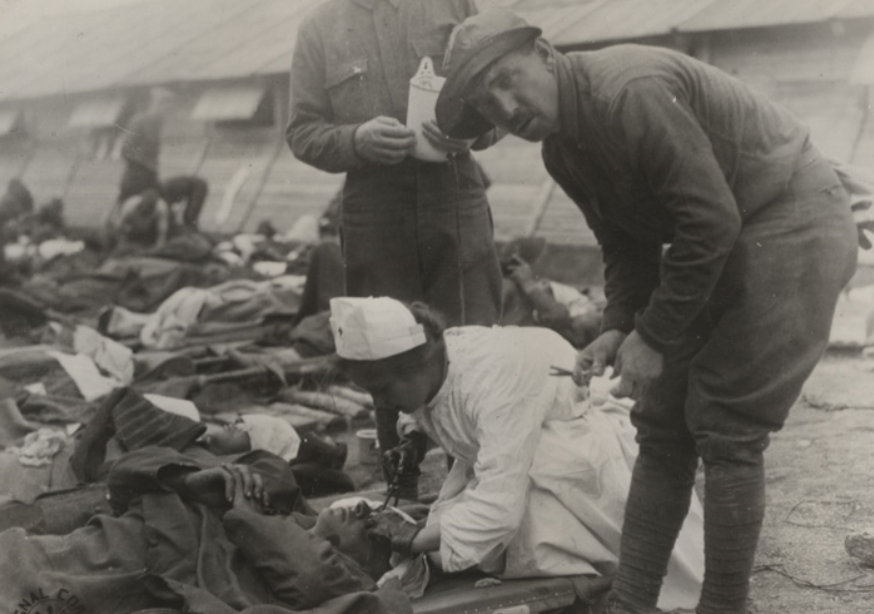 Photo of a nurse cleaning eyes of a soldier laying on a stretcher, on the ground, in the open air at a field hospital.