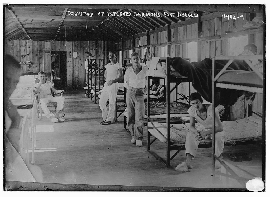 Black and white photo of a dormitory with bunk beds. Men dressed in white shirts and pants are standing around or sitting on the bunk beds