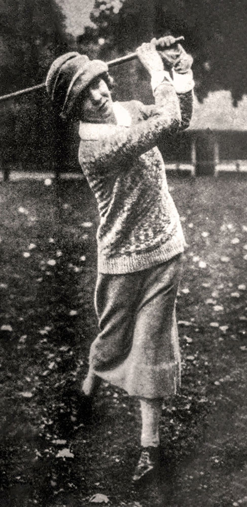 Black and white photo of a thin white woman wearing a light sweater, pencil skirt and cloche hat in mid-swing of a golf club