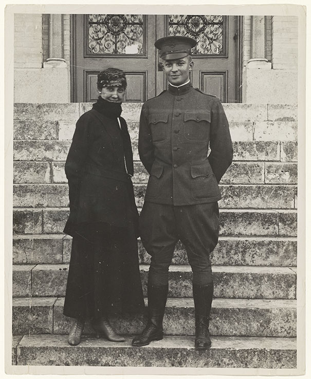 Black and white photo of a young white man in WWI military uniform and a young white woman bundled up in a dark skirt and jacket posing on the front steps leading up to some large building