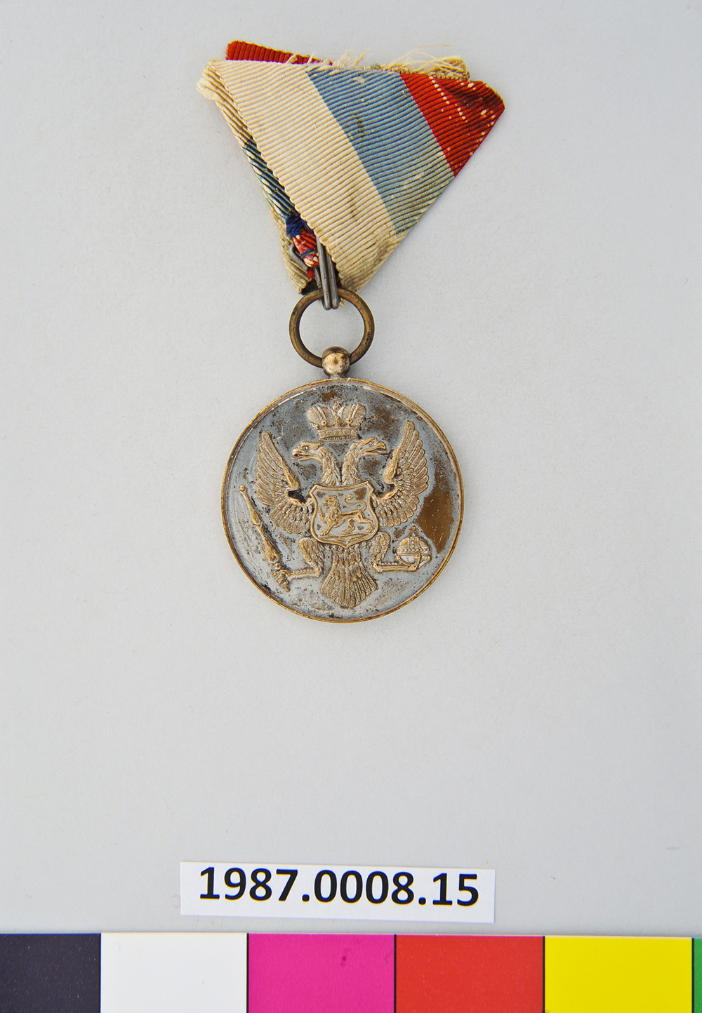 Modern photograph of a WWI-era medal hanging from a bit of ribbon. The medal is engraved with a two-headed eagle, a crown and other symbols