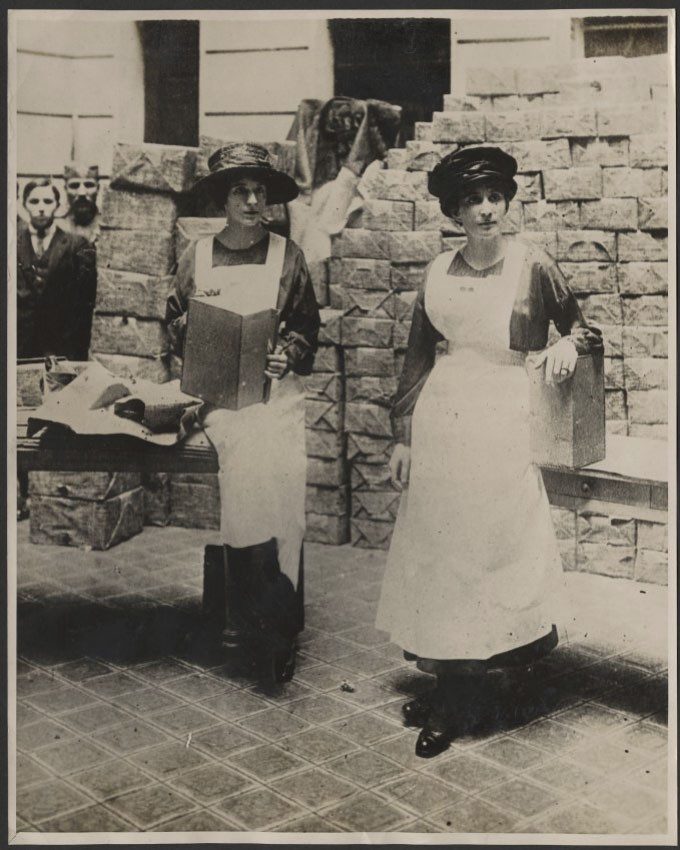 Black and white photograph of two young white women in dresses and aprons standing at tables outside a building