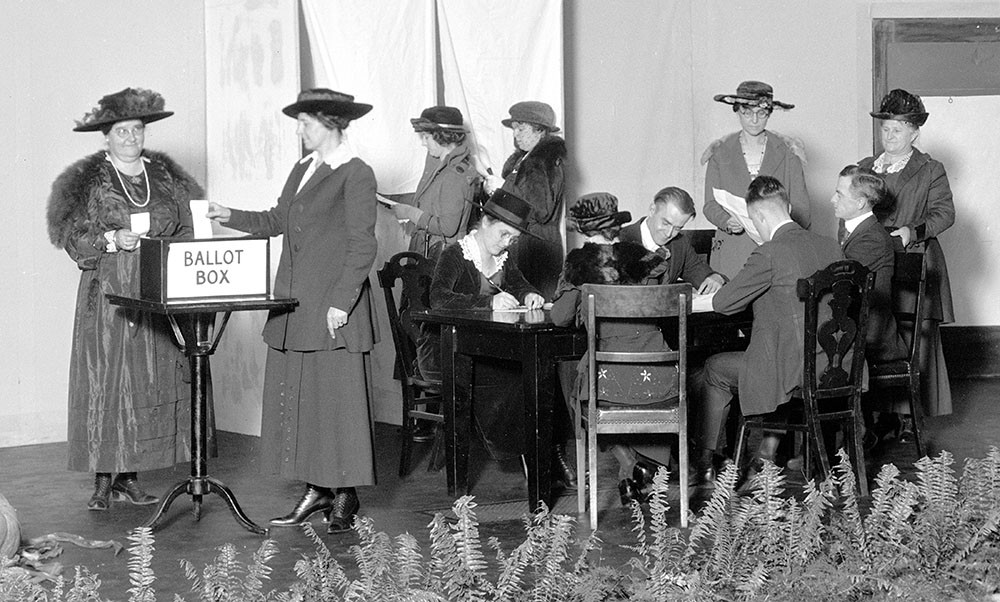 Black and white photo of a group of white women. Some are seated at a table filling out ballots, some are putting ballots into a box labeled 'Ballot Box', and some are waiting in line.