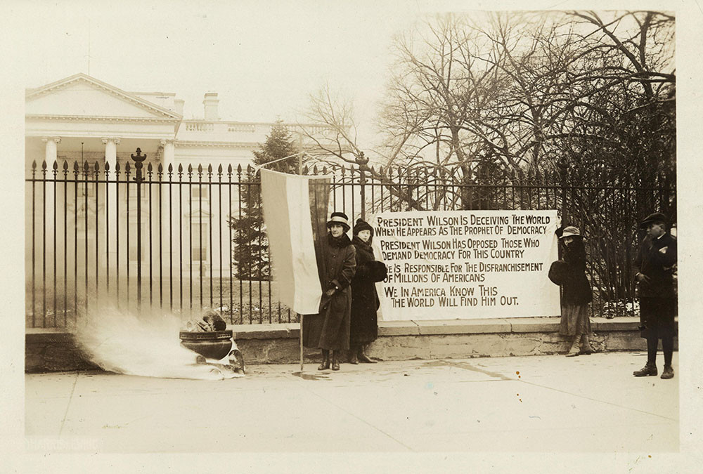 Black and white photograph of a small group of women bundled up against the cold, holding a large protest sign in front of the fence around the U.S. White House. 