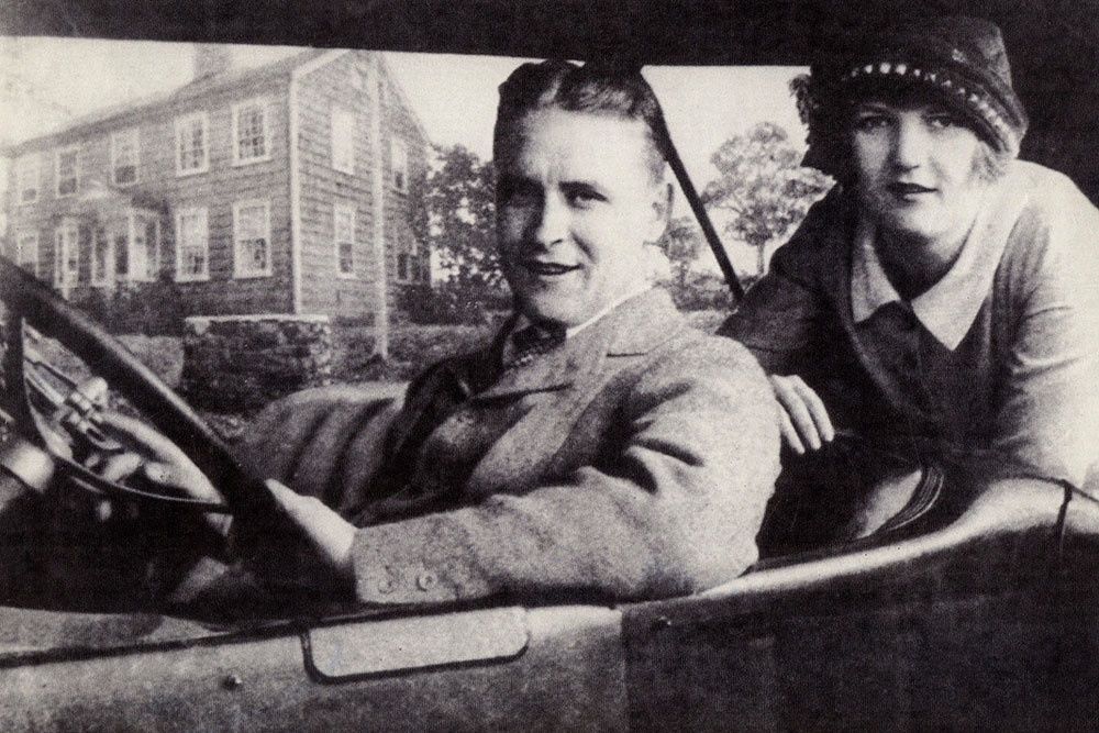 Black and white photo of a young white couple in 1920s clothing - the woman wearing a cloche hat - seated in an automobile