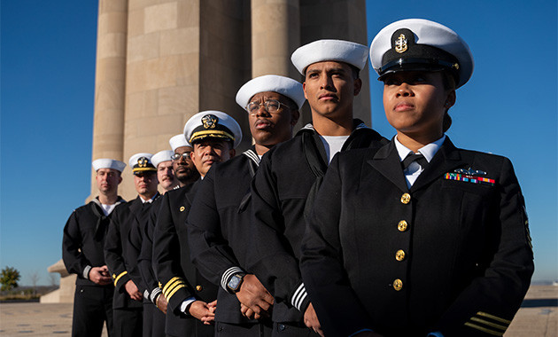 Eight U.S. Navy personnel in formal uniform standing in a row with the Liberty Memorial Tower in the background