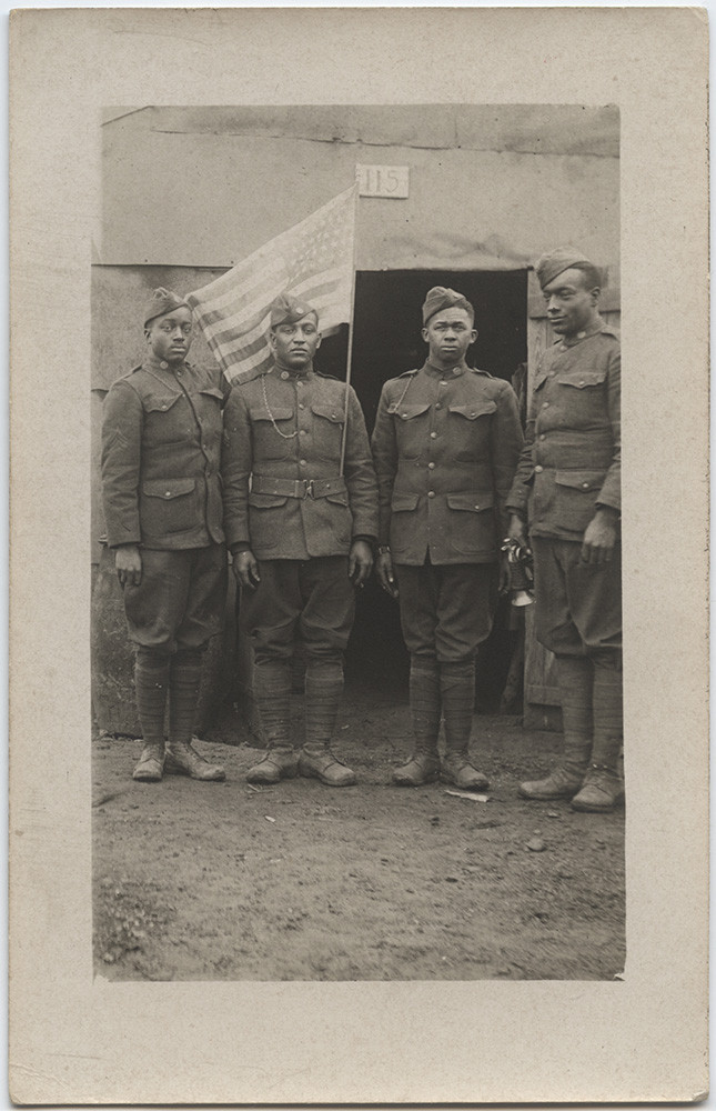Black and white postcard photograph of four African American soldiers posing. One has an American flag tucked into his belt. Soldier on right is holding a bugle in his right hand.