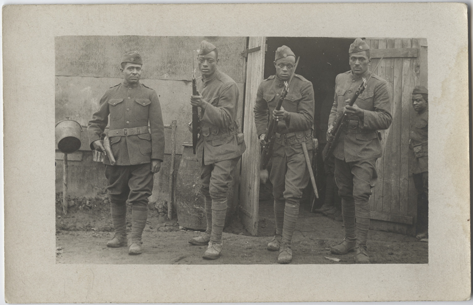  	 Black and white postcard photograph of four African American soldiers posing with their firearms with attached bayonets. They are posing in front of their barracks. 
