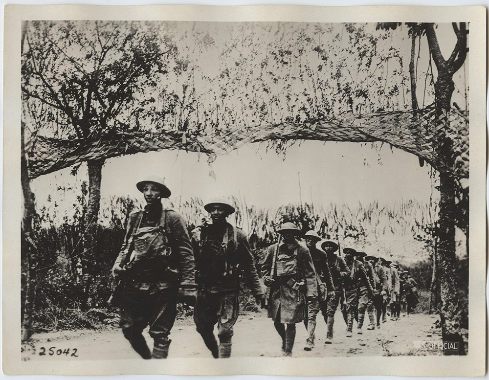 Black and white photograph of African American soldiers marching along a road that has been camouflaged. 