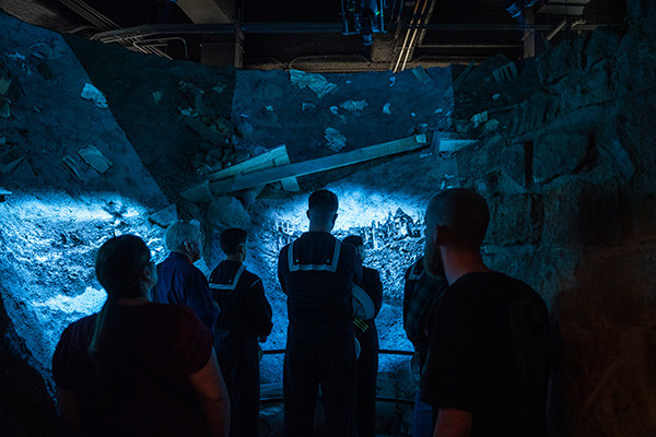 Museum guests stand inside a replica of a devastating bomb crater bathed in blue light as films are projected on the sides of the crater