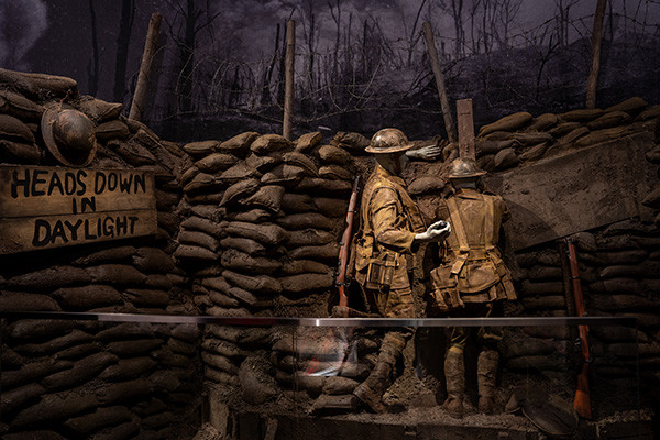 Museum exhibit replicating a British WWI trench with sandbags and wooden walkways. A soldier mannequin peers through a trench periscope while another soldier mannequin readies to throw a grenade.