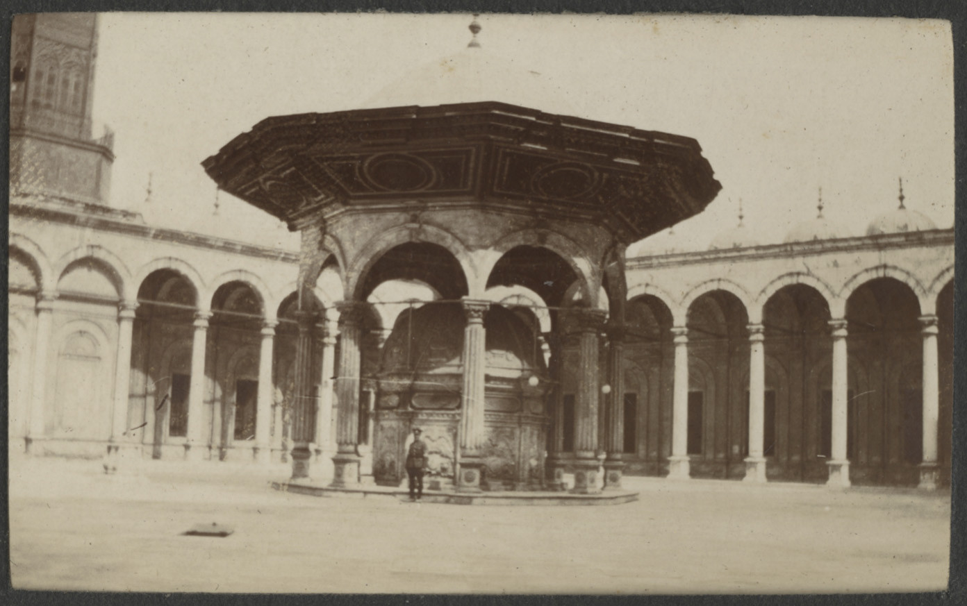 Black and white photograph of a British soldier in uniform standing under a pavilion in a courtyard