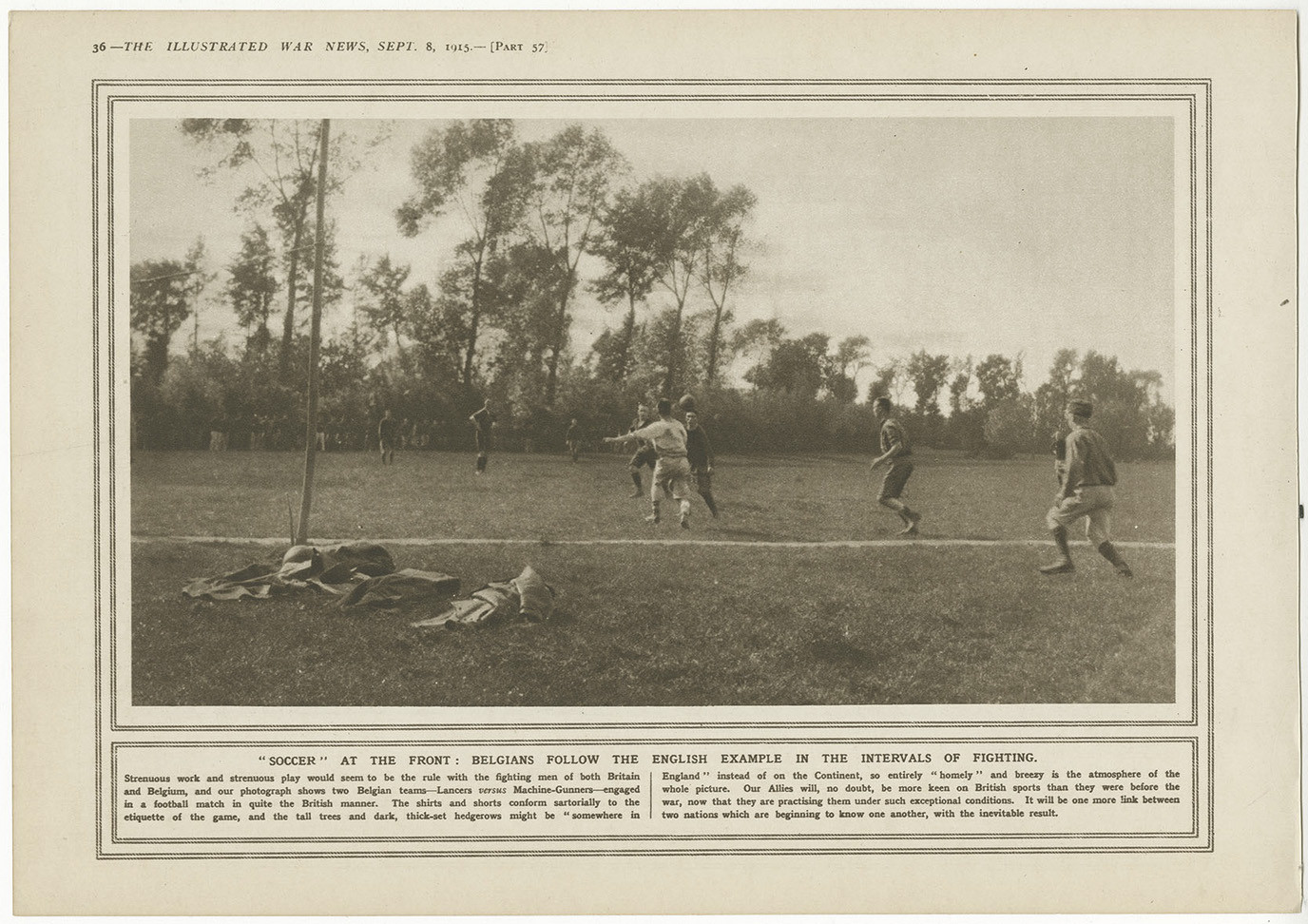 Black and white photo of WWI soldiers stripped down to shirtsleeves playing a game of football in a field surrounded by trees. Abandoned clothes and equipment lie nearby under a tree.
