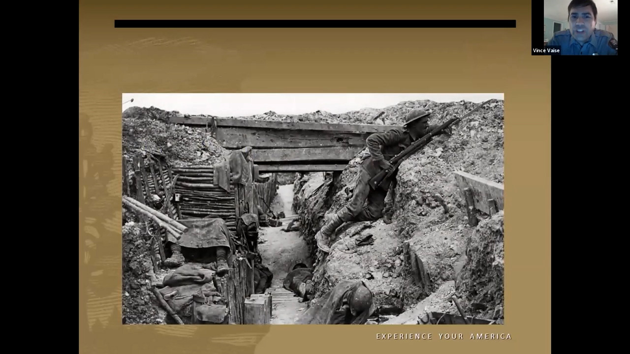 Black and white photo of a WWI soldier peering over the top of a trench with his gun at the ready