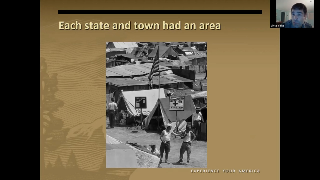 Black and white photograph of a shantytown. A tent in the foreground has a U.S. flag and a sign that saids First Aid.