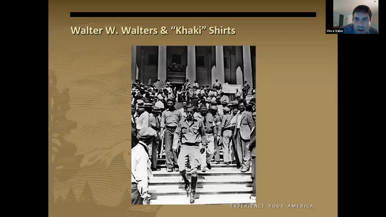 Black and white photo of a slender white man wearing tall boots, khaki pants and a khaki shirt descending the steps of a building followed by men dressed similarly