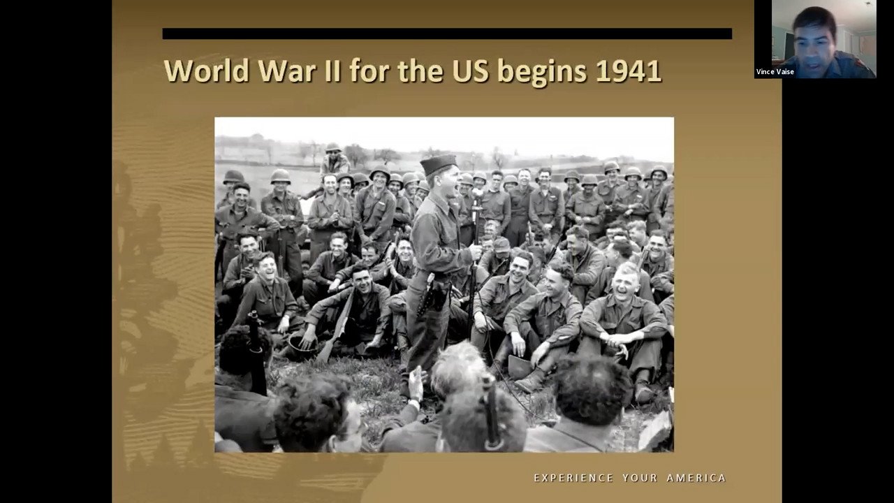 WWII soldiers sitting down in a grassy field listening to another soldier speaking into a microphone