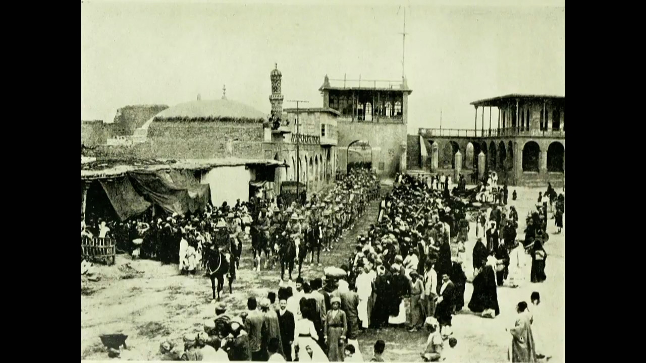 Black and white photograph of a column of horsed soldiers marching down a street while civilians look on