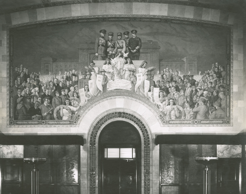 Black and white photograph of a paiting of five generals standing at a podium. Around them are a variety of different men and women. Behind them there is a large building. This mural is on a wall above a decorated door and frame and a wall with words on it.