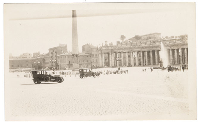 Black and white photo of a large cobblestoned square with stone palaces in the background and automobiles and tourists in the foreground