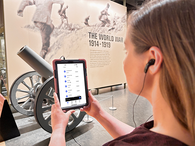 A woman holds a computer tablet up, showing the audio guide screen, as she listens on headphones