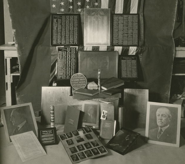 Black and white photograph of objects chosen for the Liberty Memorial Association's time capsule. Included are a series of blocks for newspaper prints, photographs of important people, an American flag, and a variety of containers.