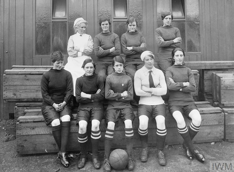 A black and white photograph with five women sitting in the foreground and four women standing behind them. The women are all wearing the same over the knee socks and there is a soccerball at the front center of the image.