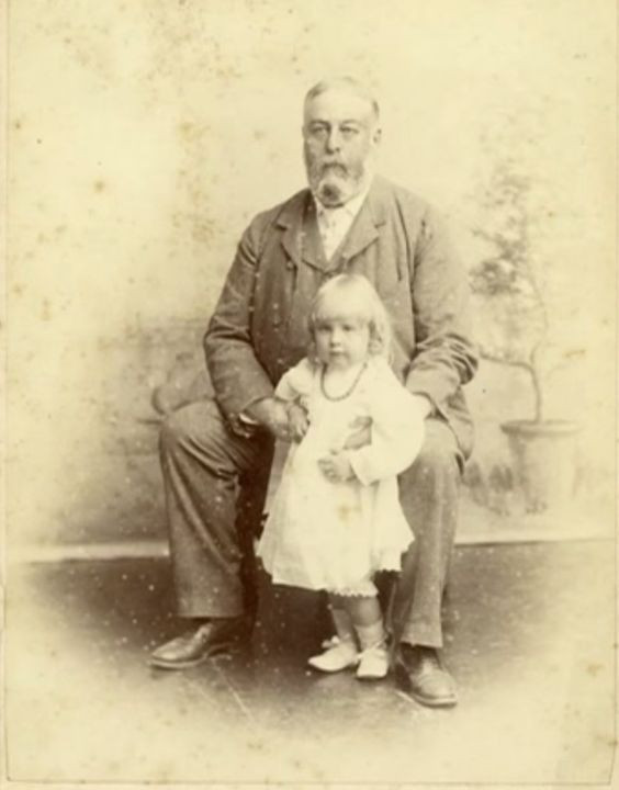 Faded sepia photo of a bearded white man in a suit sitting down for a portrait while holding a small white girl in a light-colored dress